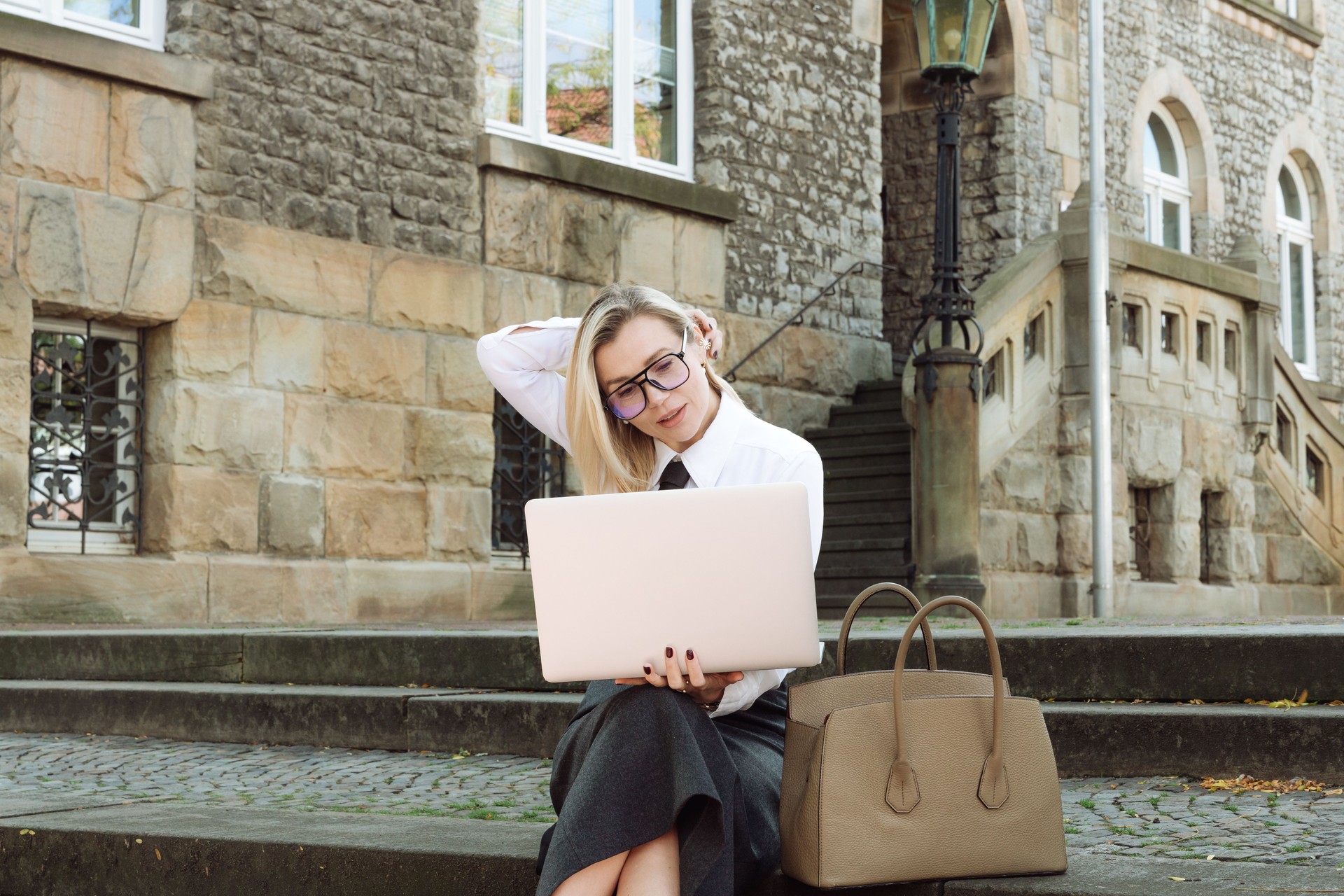 young blonde woman in business clothes sits on the steps with a laptop on the street. high quality photo